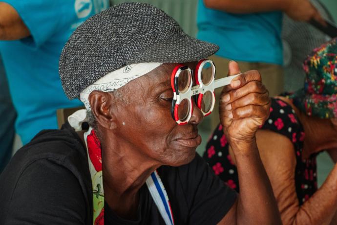 Woman looks through an eye tester while crewmembers support others in the background.