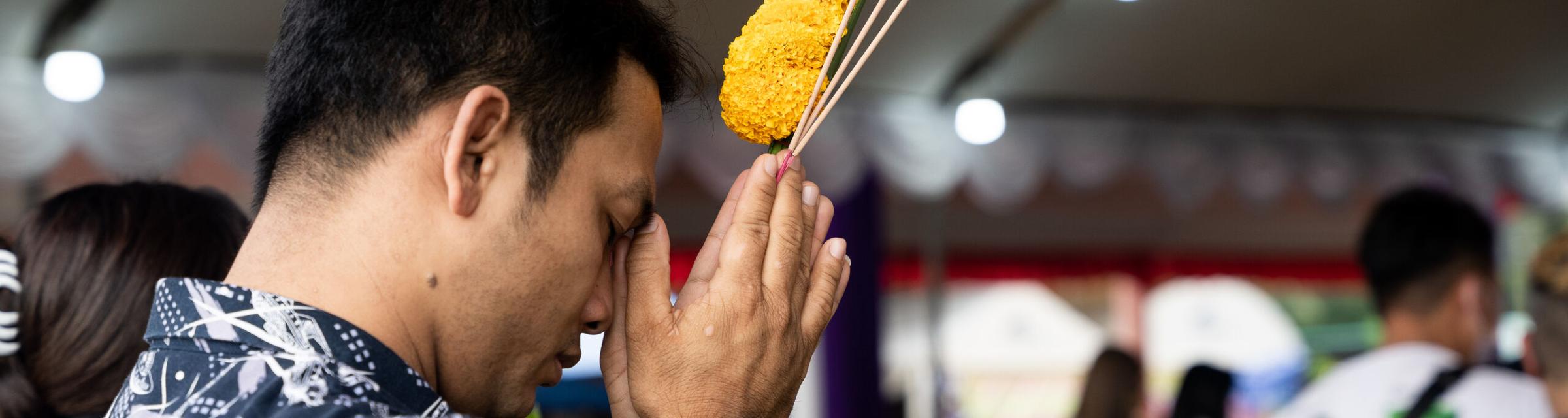 Man holding yellow flowers prays at a Buddhist shrine in Thailand. Photo by RJ Rempel.