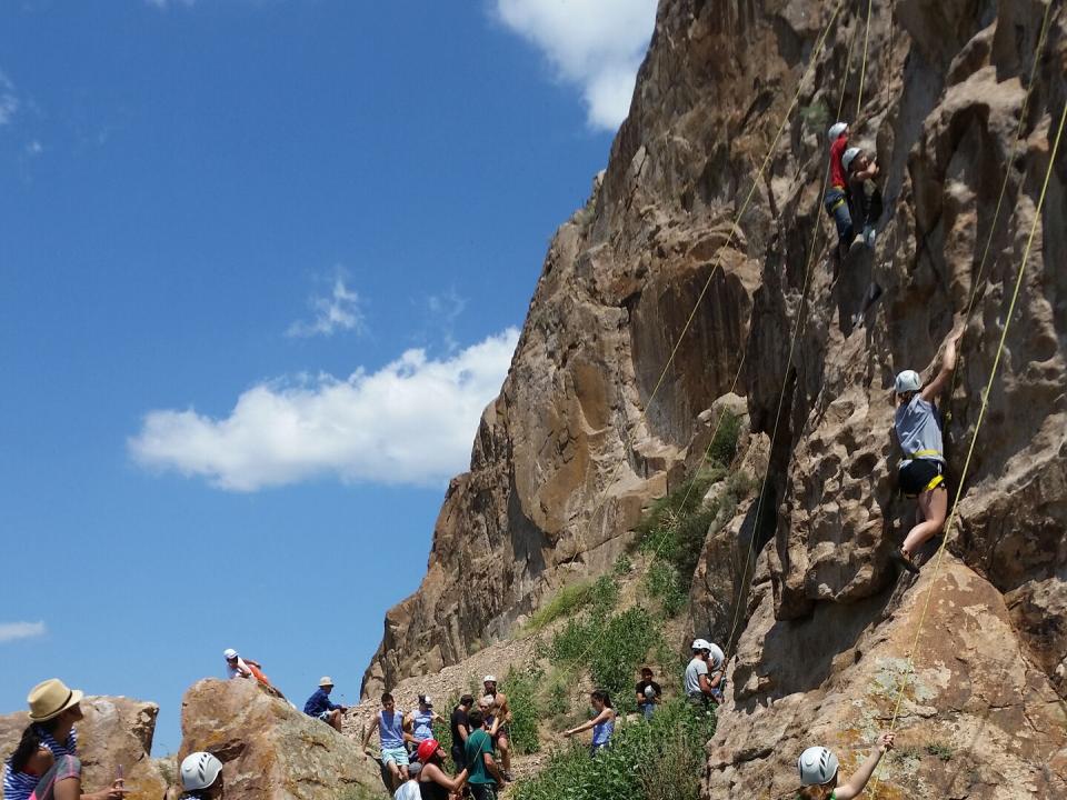 Several people climb a rock face with helmets and ropes as others look on.