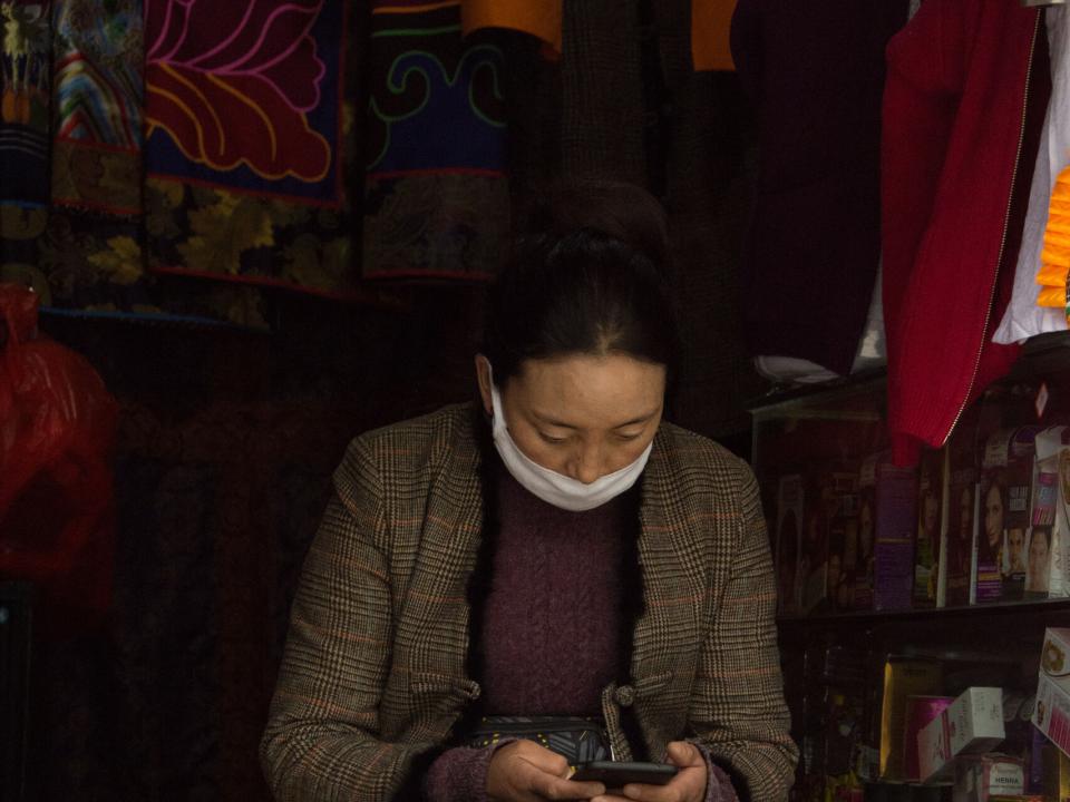 A Tibetan woman scrolls on her phone in a marketplace where she works. Photo by Ellyn Schellenberg.
