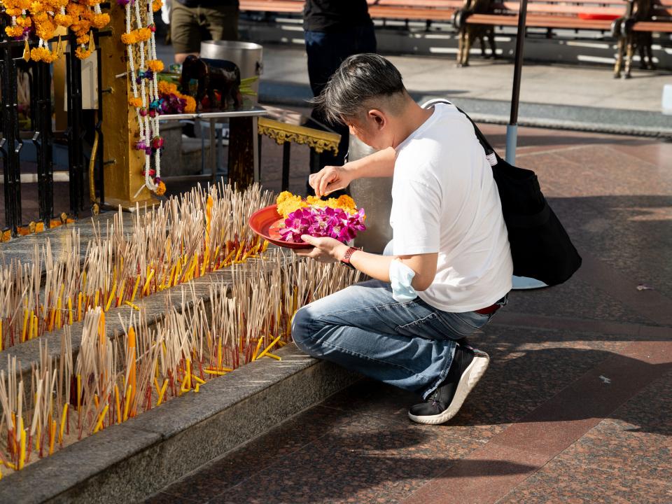 Man praying at a Buddhist shrine in Thailand holding a plate of purple and yellow flowers. Photo by Rebecca Rempel.