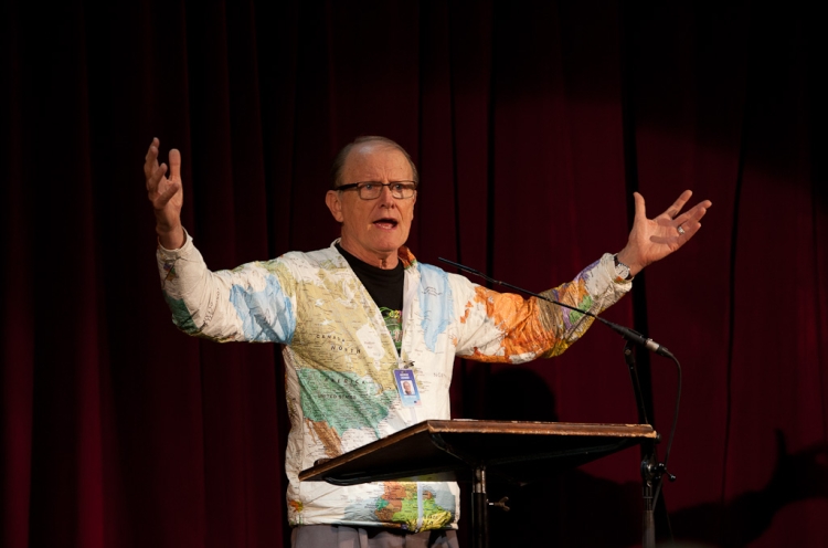 George Verwer predigt mit ausgebreiteten Armen und Weltkarten-Jacke