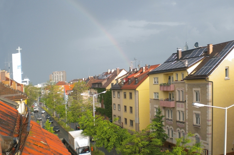 Regenbogen über den Dächern einer Stadt. Links im Bild ist ein Kirchturm mit Kreuz